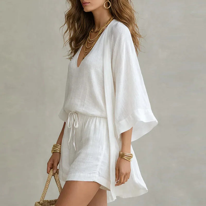 White - A woman wearing a white three-piece linen set featuring a V-neck tank top, high-waisted drawstring shorts, and a matching long open-front duster cardigan, accessorized with gold layered necklaces and a woven straw bag.
