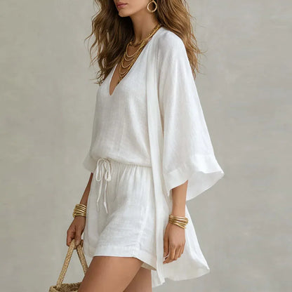 White - A woman wearing a white three-piece linen set featuring a V-neck tank top, high-waisted drawstring shorts, and a matching long open-front duster cardigan, accessorized with gold layered necklaces and a woven straw bag.