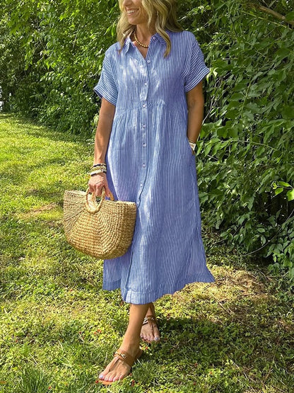 Blue - A woman wearing a blue and white vertically striped linen-blend midi shirt dress with short sleeves, a pointed collar, and a button-down front, holding a woven straw bag in a grassy outdoor setting.