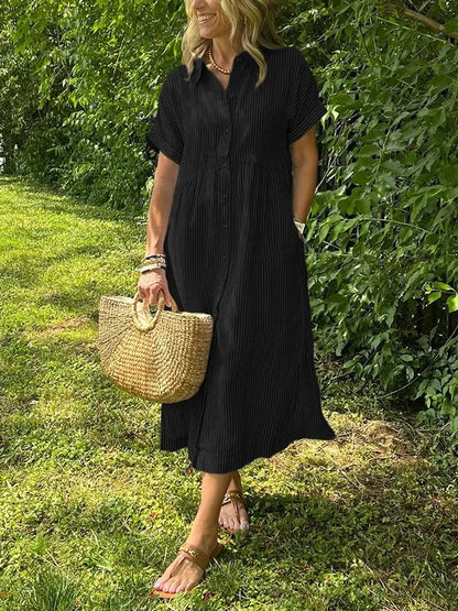 Black - A woman wearing a blue and white vertically striped linen-blend midi shirt dress with short sleeves, a pointed collar, and a button-down front, holding a woven straw bag in a grassy outdoor setting.