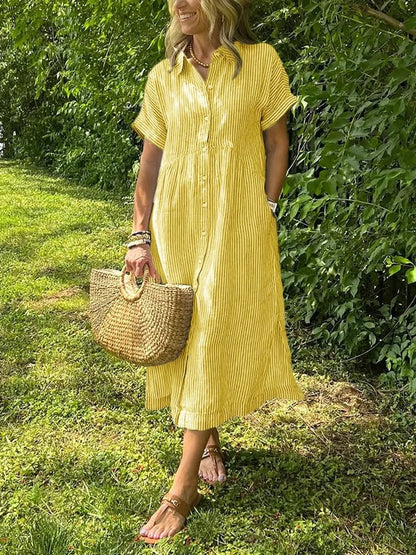 Yellow - A woman wearing a blue and white vertically striped linen-blend midi shirt dress with short sleeves, a pointed collar, and a button-down front, holding a woven straw bag in a grassy outdoor setting.