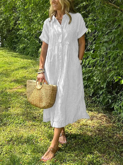 White - A woman wearing a blue and white vertically striped linen-blend midi shirt dress with short sleeves, a pointed collar, and a button-down front, holding a woven straw bag in a grassy outdoor setting.