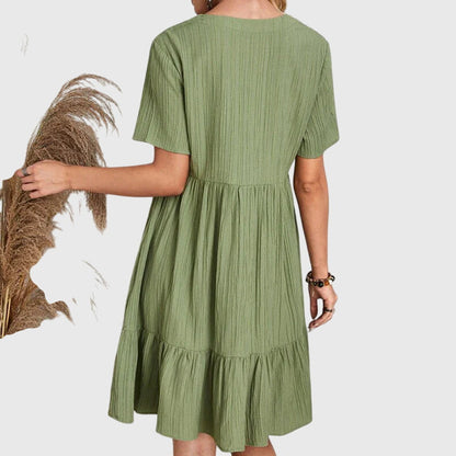Pink - A woman wearing a dusty rose crinkle gauze short-sleeve V-neck sundress with an empire waist and tiered ruffle hem, styled with a straw shoulder bag.