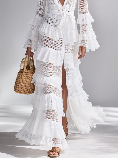 White - Woman wearing a crochet tiered maxi dress with long bell sleeves, a plunging neckline, and a waist tie, holding a woven basket bag