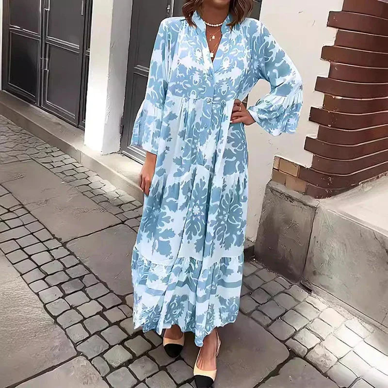 Blue - A woman wearing a cream and tan damask print maxi sundress with long bell sleeves, a V-neckline, and a tiered skirt, standing on a cobblestone street.