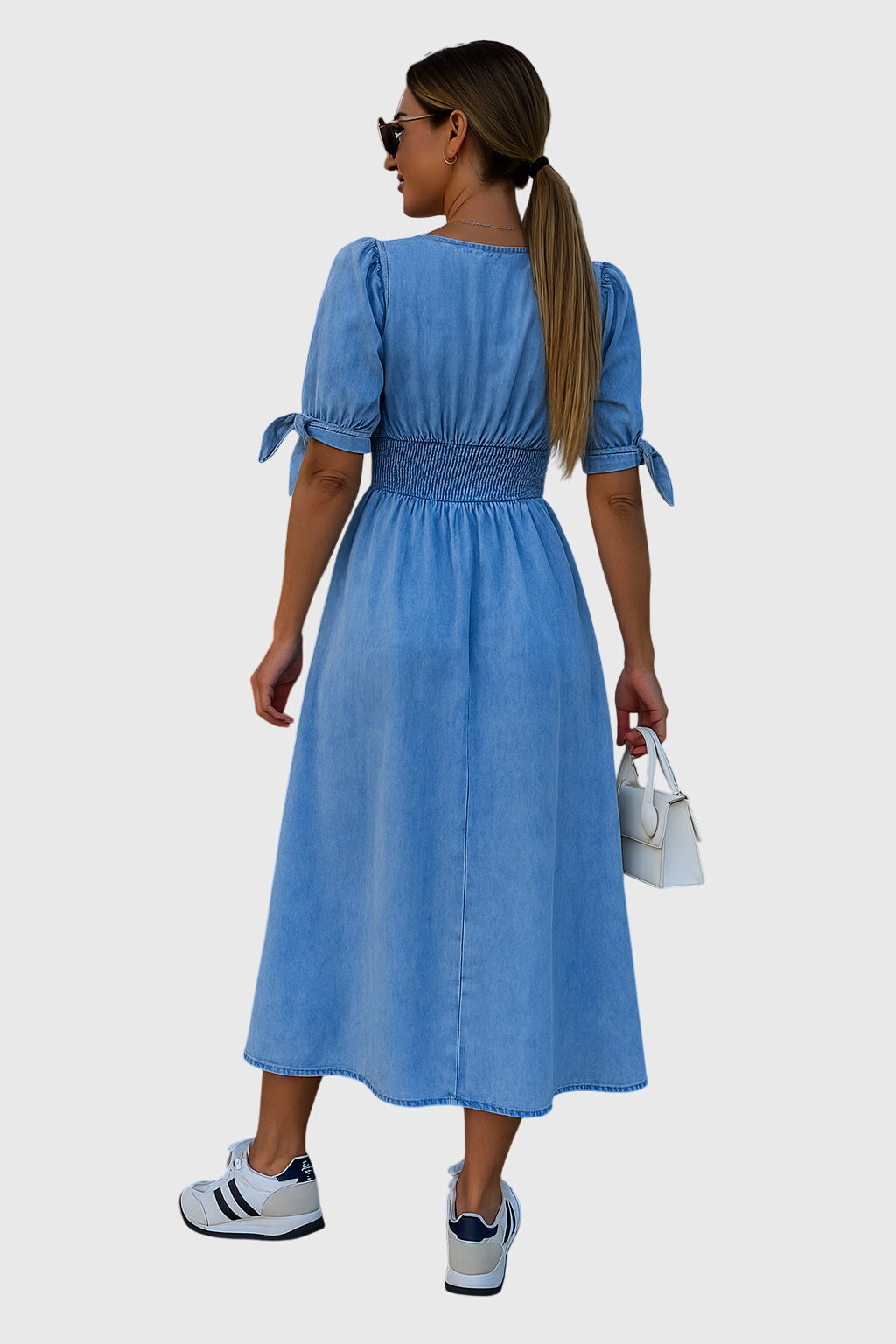 Blue - A woman wearing a light-wash blue denim midi dress with puff sleeves, tie cuffs, a V-neckline, and a button-down front, styled with white sneakers and a small white handbag.