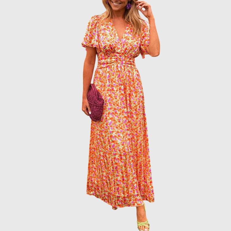 A woman wearing an orange and pink ditsy floral print maxi dress with a V-neckline, flutter sleeves, and a ruched empire waist.