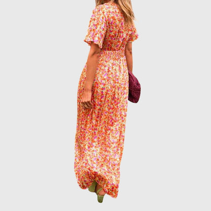 A woman wearing an orange and pink ditsy floral print maxi dress with a V-neckline, flutter sleeves, and a ruched empire waist.