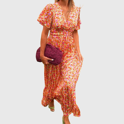 A woman wearing an orange and pink ditsy floral print maxi dress with a V-neckline, flutter sleeves, and a ruched empire waist.
