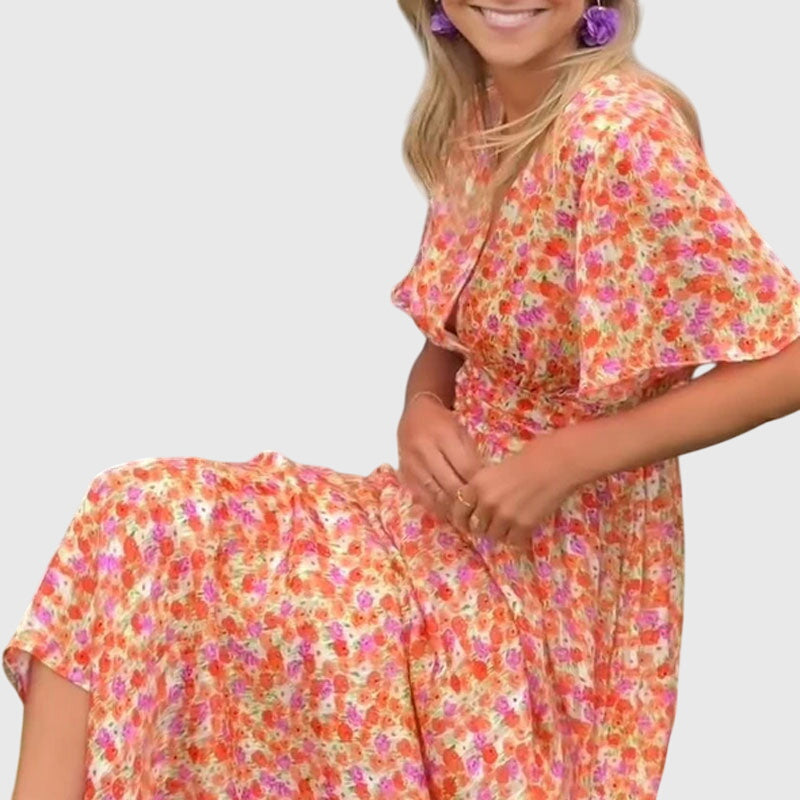 A woman wearing an orange and pink ditsy floral print maxi dress with a V-neckline, flutter sleeves, and a ruched empire waist.