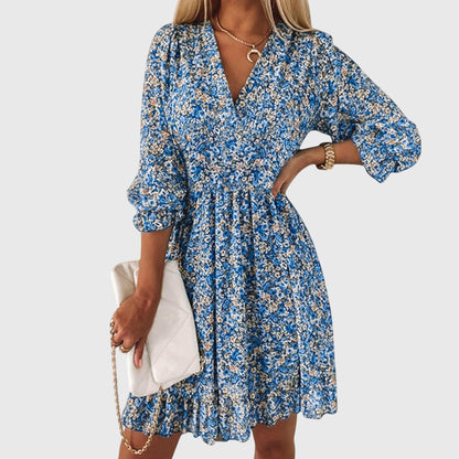 Blue - A woman wearing a blue and white ditsy floral print sundress with a v-neckline, three-quarter sleeves, a cinched waist, and a ruffled hem, holding a white quilted clutch bag.