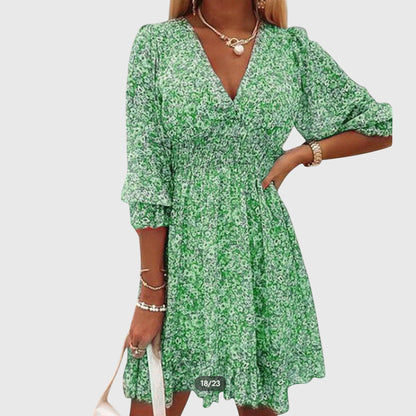 Green - A woman wearing a blue and white ditsy floral print sundress with a v-neckline, three-quarter sleeves, a cinched waist, and a ruffled hem, holding a white quilted clutch bag.