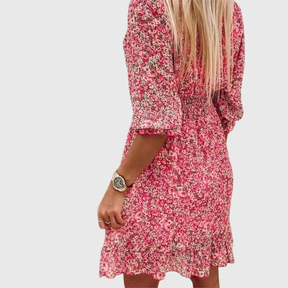 Pink - A woman wearing a blue and white ditsy floral print sundress with a v-neckline, three-quarter sleeves, a cinched waist, and a ruffled hem, holding a white quilted clutch bag.