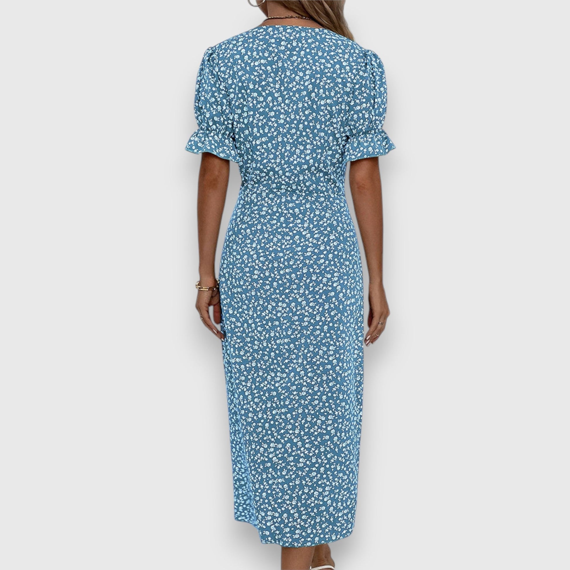 A woman wearing a light blue midi sundress with a white ditsy floral print, featuring puff sleeves, a V-neckline, and an asymmetrical wrap skirt with button details.