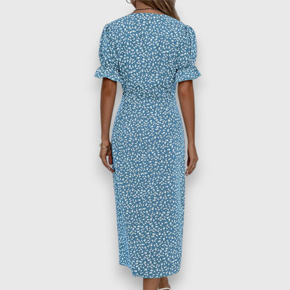 A woman wearing a light blue midi sundress with a white ditsy floral print, featuring puff sleeves, a V-neckline, and an asymmetrical wrap skirt with button details.