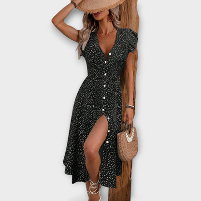 Model wearing a red and white ditsy polka dot midi sundress with a V-neckline, flutter sleeves, and button-down front, paired with a straw hat and white lace-up heels.