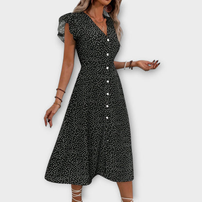 Model wearing a red and white ditsy polka dot midi sundress with a V-neckline, flutter sleeves, and button-down front, paired with a straw hat and white lace-up heels.