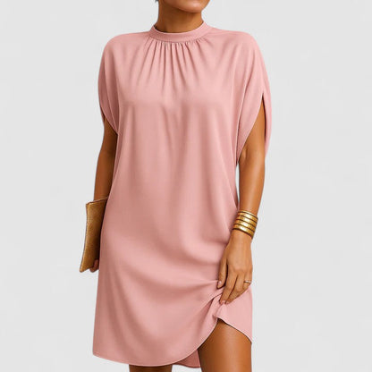 Pink - A woman wearing a dusty pink crepe shift dress featuring a gathered mock neckline, draped cap sleeves, and a relaxed silhouette.