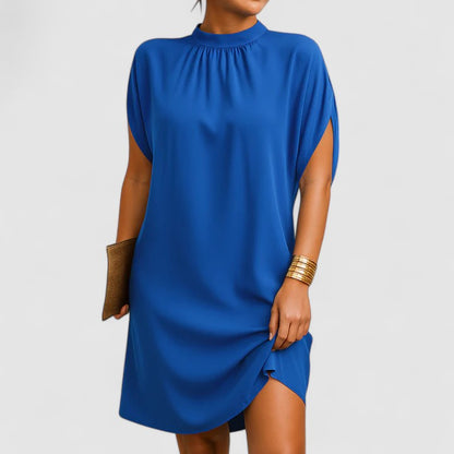 Blue - A woman wearing a dusty pink crepe shift dress featuring a gathered mock neckline, draped cap sleeves, and a relaxed silhouette.