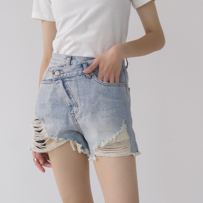 Woman wearing light wash denim shorts with asymmetrical waist and distressed hem.