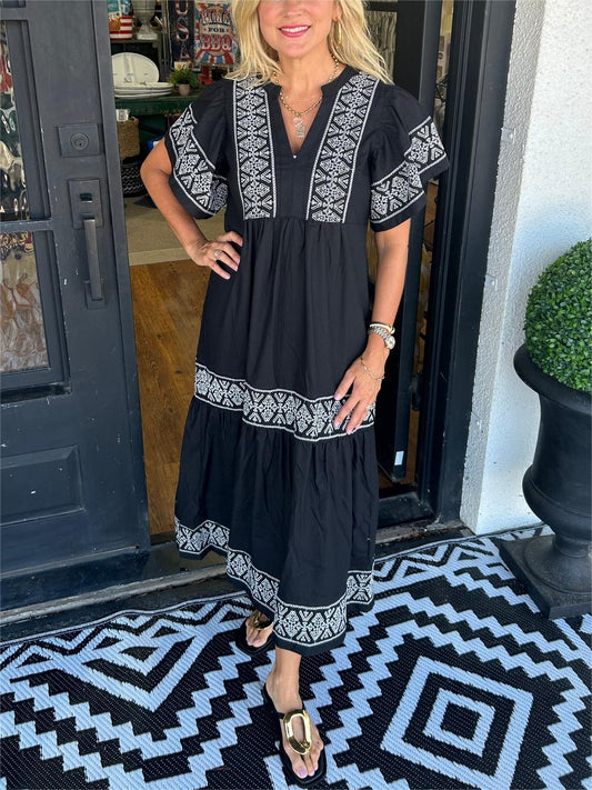 Black - A woman wearing a black midi-length cotton dress featuring white geometric embroidery along the V-neckline, flutter sleeves, tiered skirt, and hem.