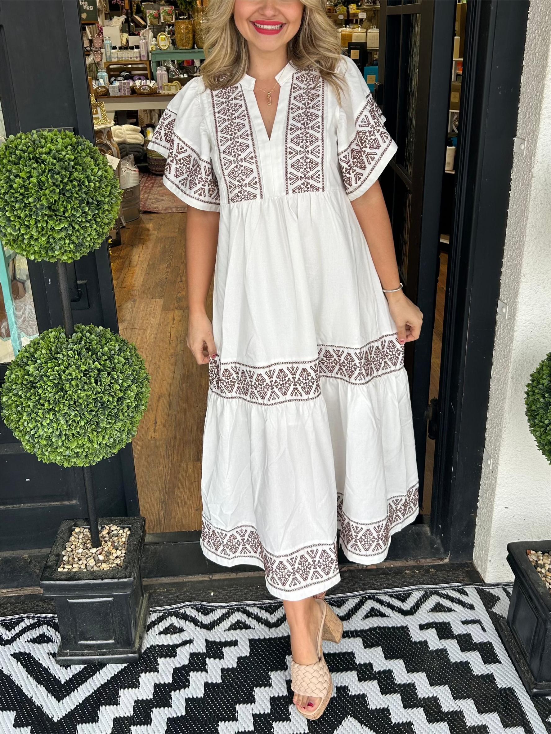 White - A woman wearing a black midi-length cotton dress featuring white geometric embroidery along the V-neckline, flutter sleeves, tiered skirt, and hem.