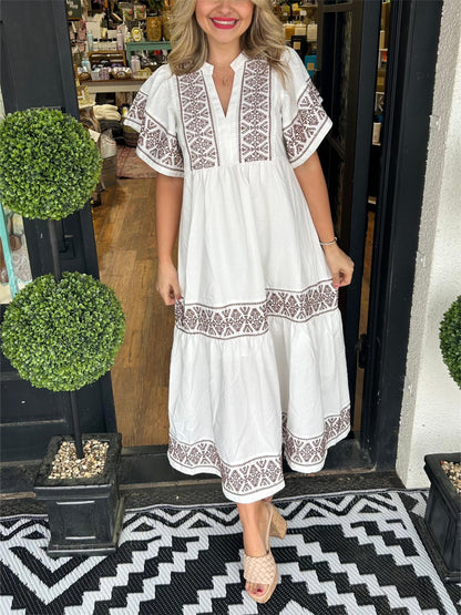 White - A woman wearing a black midi-length cotton dress featuring white geometric embroidery along the V-neckline, flutter sleeves, tiered skirt, and hem.