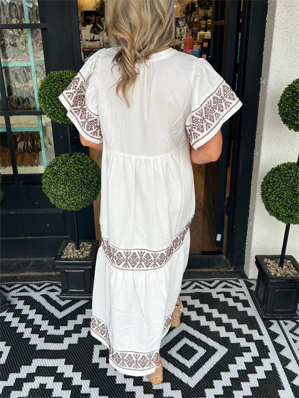 White - A woman wearing a black midi-length cotton dress featuring white geometric embroidery along the V-neckline, flutter sleeves, tiered skirt, and hem.