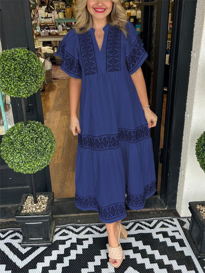 Navy Blue - A woman wearing a black midi-length cotton dress featuring white geometric embroidery along the V-neckline, flutter sleeves, tiered skirt, and hem.