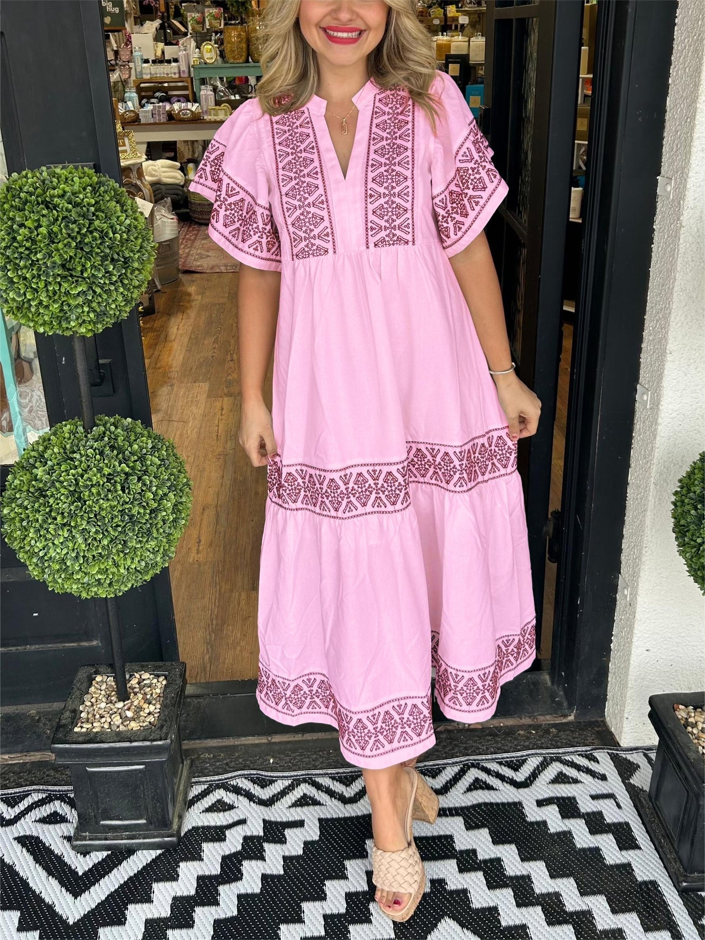 Pink - A woman wearing a black midi-length cotton dress featuring white geometric embroidery along the V-neckline, flutter sleeves, tiered skirt, and hem.
