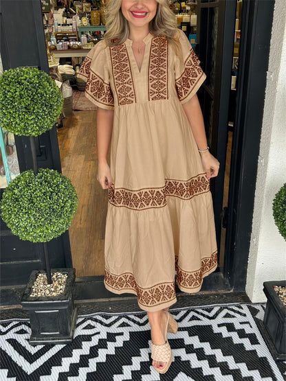 Mocha - A woman wearing a black midi-length cotton dress featuring white geometric embroidery along the V-neckline, flutter sleeves, tiered skirt, and hem.