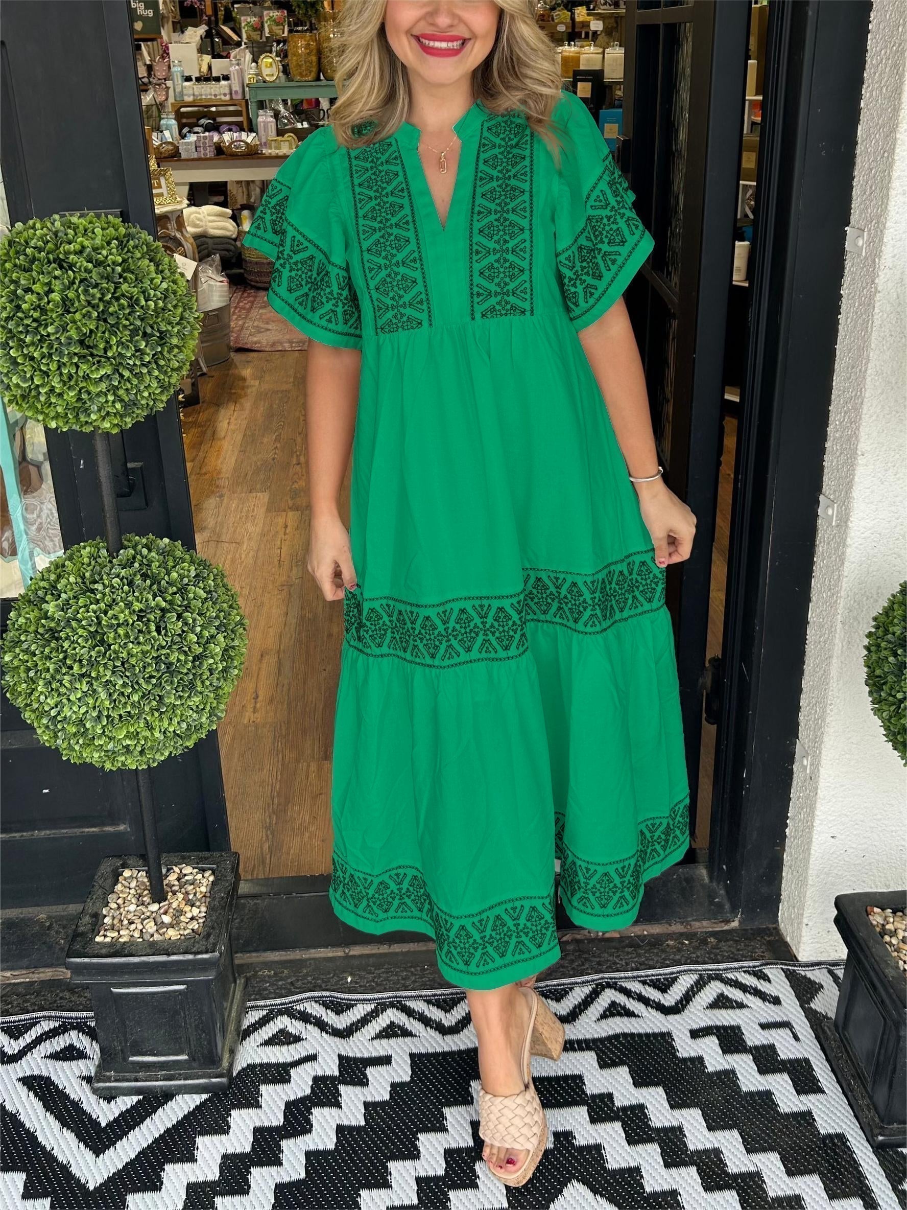 Green - A woman wearing a black midi-length cotton dress featuring white geometric embroidery along the V-neckline, flutter sleeves, tiered skirt, and hem.