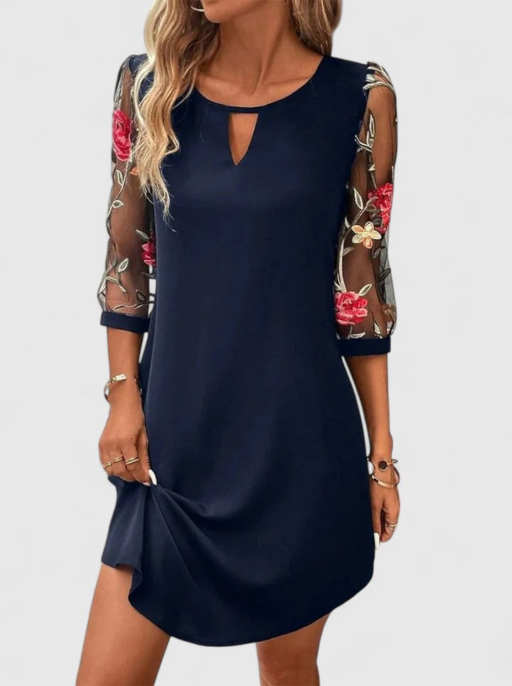 Navy Blue - A woman wearing a navy blue shift mini dress featuring sheer 3/4-length mesh sleeves with red and green floral embroidery and a small keyhole cutout at the neckline.