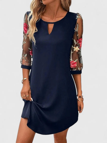 Navy Blue - A woman wearing a navy blue shift mini dress featuring sheer 3/4-length mesh sleeves with red and green floral embroidery and a small keyhole cutout at the neckline.