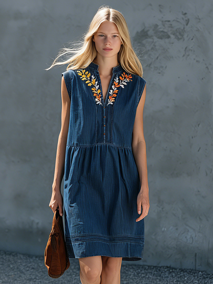 Denim - Woman walking outdoors wearing a sleeveless denim-style midi dress featuring floral embroidery around a split neckline and a relaxed gathered waist.