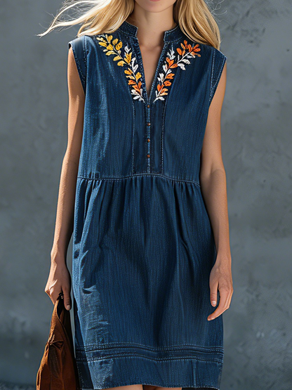Denim - Woman walking outdoors wearing a sleeveless denim-style midi dress featuring floral embroidery around a split neckline and a relaxed gathered waist.