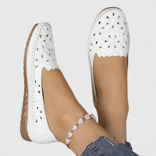 Women's slip-on loafers featuring a faux-leather upper with detailed floral cutouts, scalloped edges, and a textured flat sole.