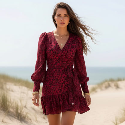 Red - A woman stands on a sandy beach wearing a black and blush pink floral print mini wrap dress with long bishop sleeves, a V-neckline, and a ruffled hem.