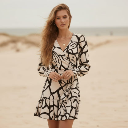 White - A woman stands on a sandy beach wearing a black and blush pink floral print mini wrap dress with long bishop sleeves, a V-neckline, and a ruffled hem.