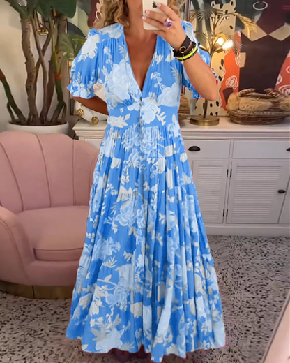 Light Blue - Woman wearing a floral print maxi dress with short puff sleeves, a v-neckline, button front, and a tiered skirt, standing with one hand in her pocket