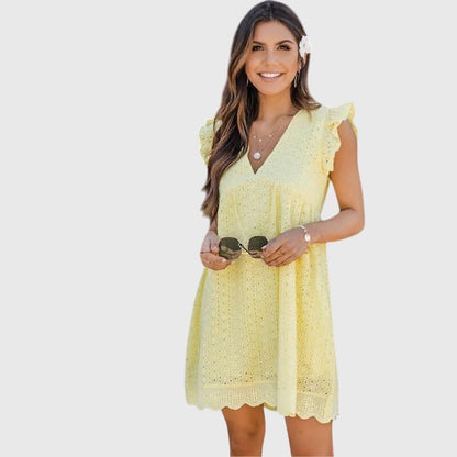 Yellow - A woman wearing a black V-neck cotton eyelet mini sundress with flutter sleeves and a scalloped hem, holding a straw clutch.