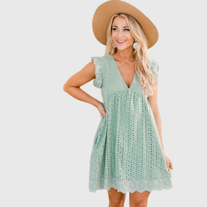 Green - A woman wearing a black V-neck cotton eyelet mini sundress with flutter sleeves and a scalloped hem, holding a straw clutch.