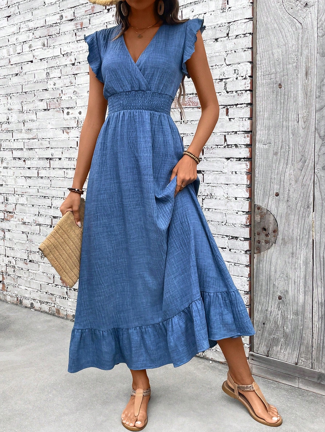 Blue - A woman wearing a dusty blue slub-textured midi sundress with flutter sleeves, a smocked waist, and a ruffled hem, paired with flat sandals and a straw clutch.