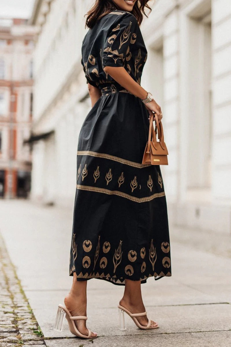 Black - A woman wearing a black midi-length shirt dress with a gold geometric border print, featuring a collared neckline, short sleeves, a belted waist, and an A-line skirt.
