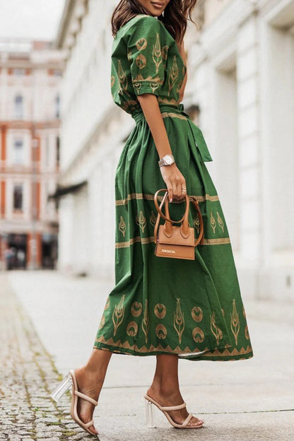 Black - A woman wearing a black midi-length shirt dress with a gold geometric border print, featuring a collared neckline, short sleeves, a belted waist, and an A-line skirt.