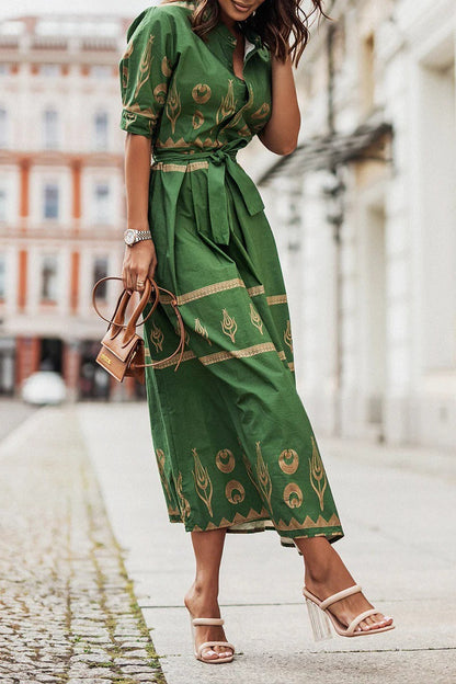 Green - A woman wearing a black midi-length shirt dress with a gold geometric border print, featuring a collared neckline, short sleeves, a belted waist, and an A-line skirt.