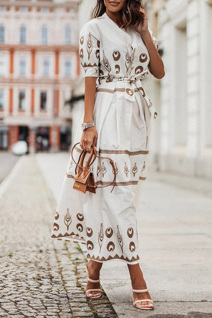 White - A woman wearing a black midi-length shirt dress with a gold geometric border print, featuring a collared neckline, short sleeves, a belted waist, and an A-line skirt.