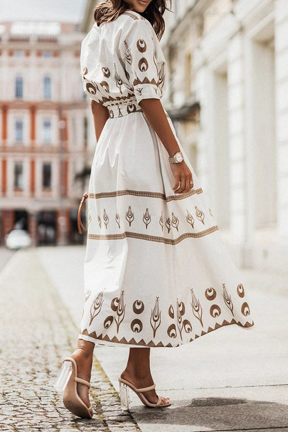 White - A woman wearing a black midi-length shirt dress with a gold geometric border print, featuring a collared neckline, short sleeves, a belted waist, and an A-line skirt.