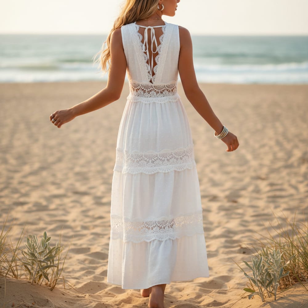 White - A woman stands on a sandy beach wearing a white sleeveless maxi dress featuring intricate lace inserts, a V-neckline, and a tiered Swiss dot skirt.