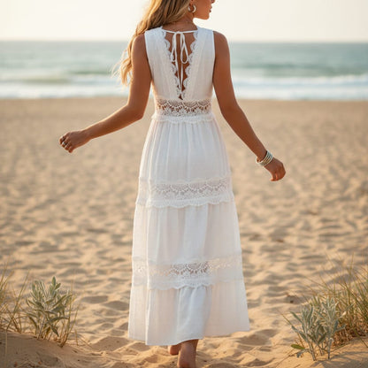 White - A woman stands on a sandy beach wearing a white sleeveless maxi dress featuring intricate lace inserts, a V-neckline, and a tiered Swiss dot skirt.